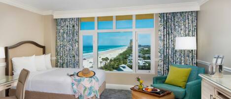 A hotel room with a bed, a desk, a chair, a sofa, a window with a beach view, and a tray with fruits and a hat.