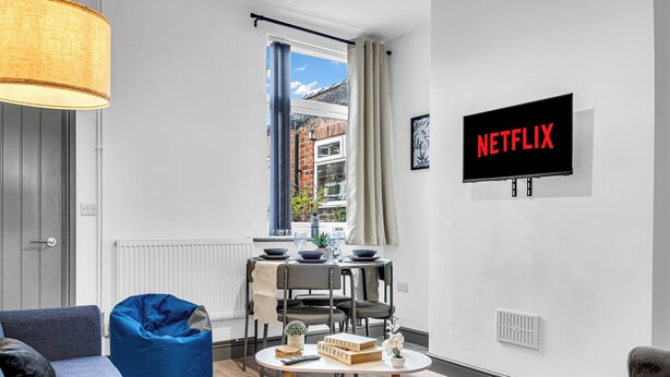 En moderne stue med spiseplads, et stort vindue med gardiner, et vægmonteret tv og et hyggeligt siddeområde.