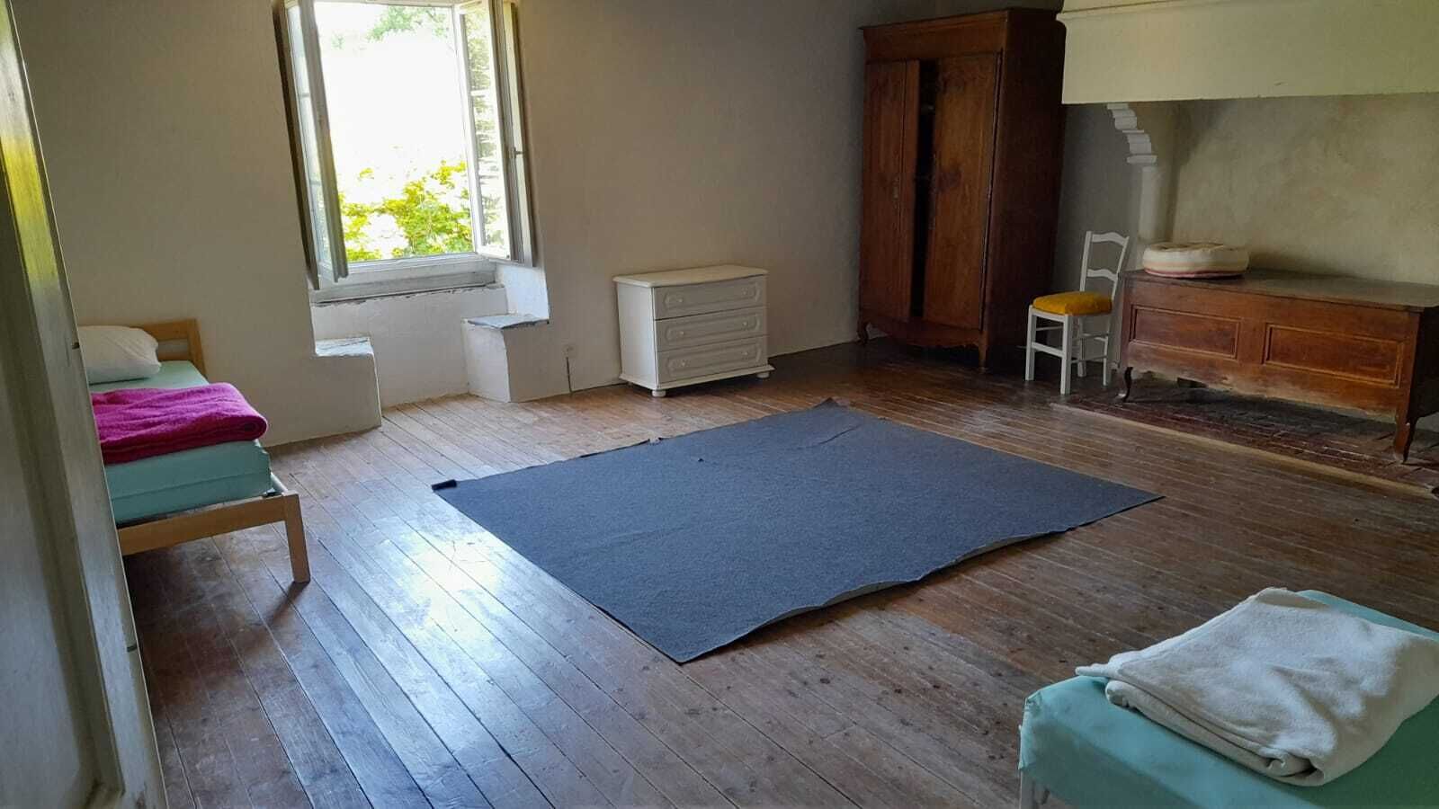 Ein Zimmer mit Holzboden, einer großen, dunklen Matte, einem Bett mit rosa Decke, einem weißen Heizkörper, einem Holzschrank und einem Fenster mit Tageslicht.