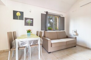 A dining area with a table, chairs, and a sofa. There are two wall-mounted air conditioning units and a window with curtains.