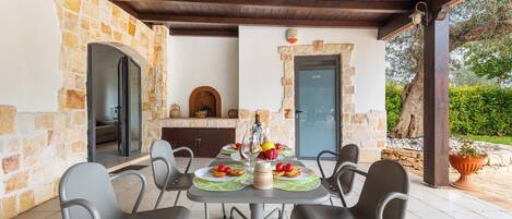 Un espace extérieur abrité, aménagé pour les repas, avec des murs en pierre, une table dressée avec des assiettes, des verres et un panier de fruits.