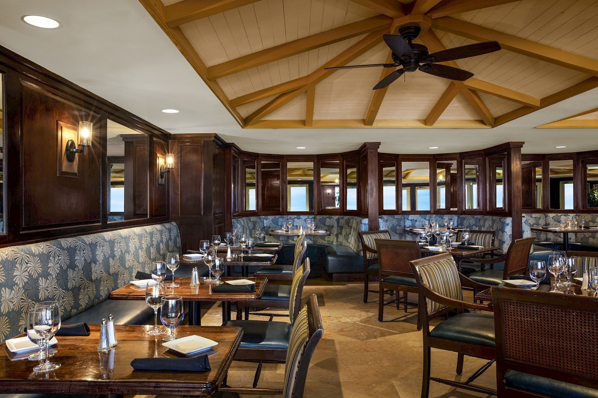 A restaurant with high wooden ceilings, a ceiling fan, and a mix of booths and tables with wine glasses set.