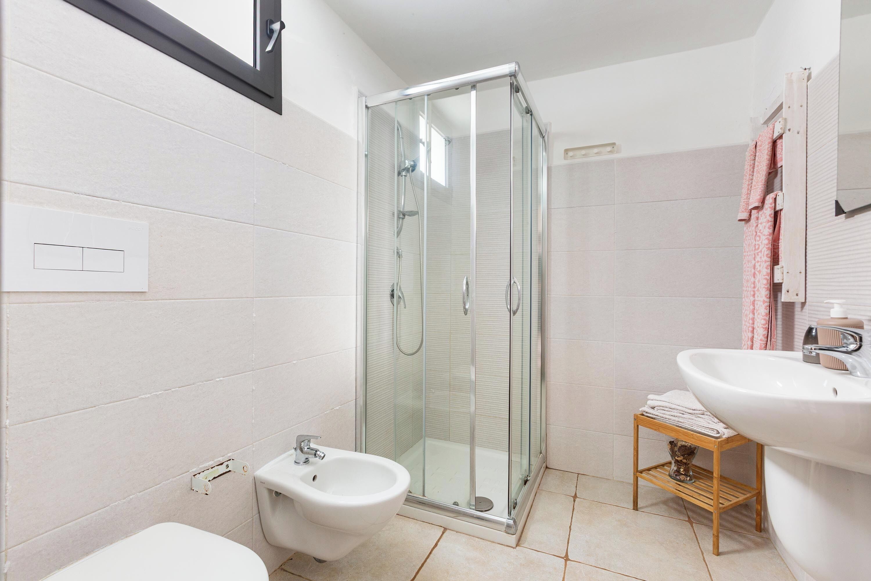A bathroom with a glass-enclosed shower, a bidet, a sink, and a small wooden shelf.