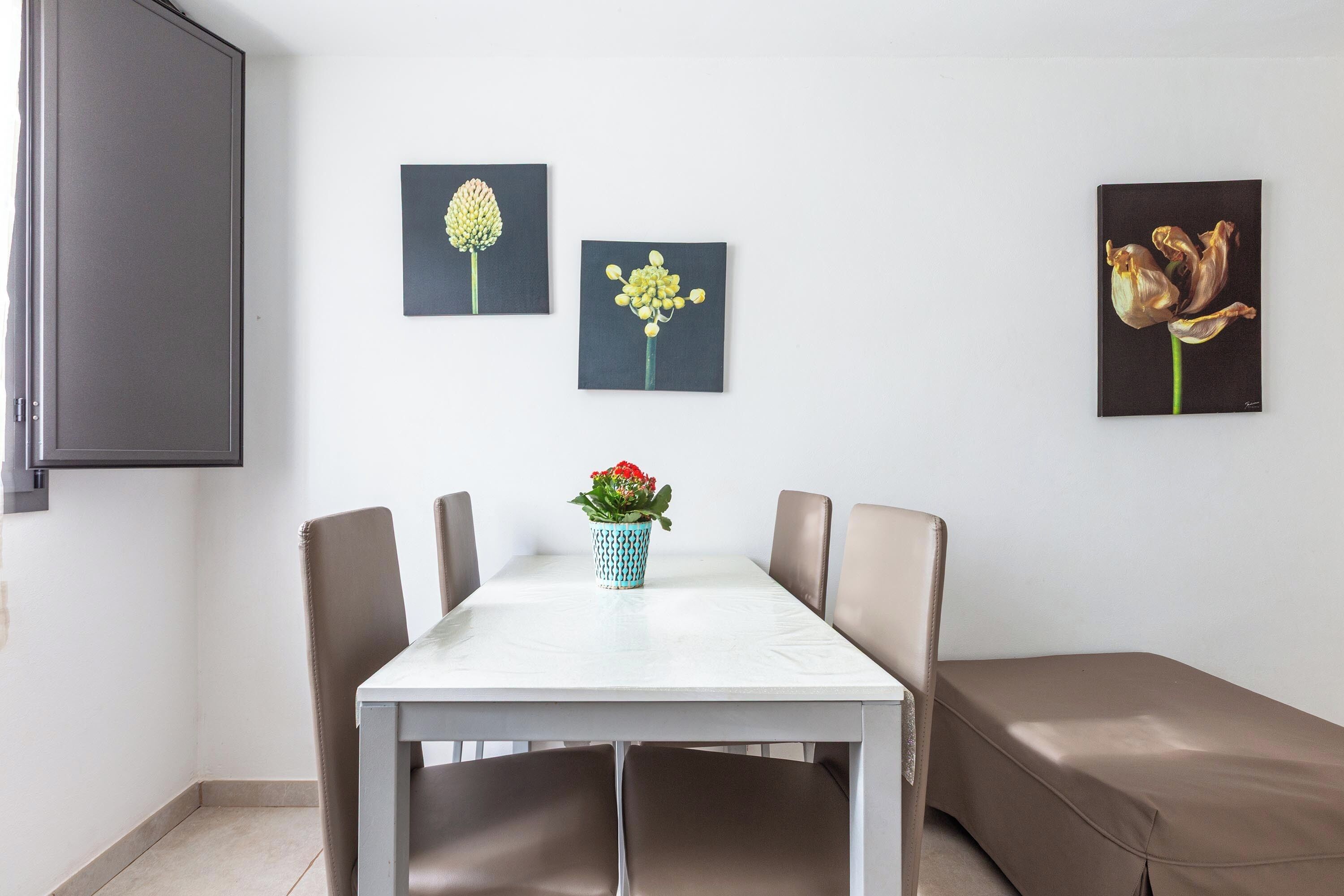 A dining area with a white table, four chairs, a small potted plant, and three framed artworks on the wall.