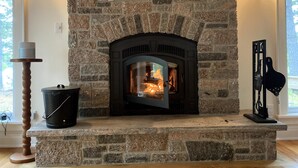 Une cheminée en pierre avec un feu de bois, un seau noir et un support en bois.