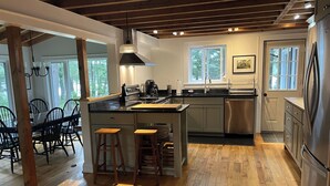 Une cuisine moderne avec un plancher en bois, un îlot central, des tabourets de bar, un réfrigérateur en acier inoxydable et une fenêtre laissant entrer la lumière naturelle.