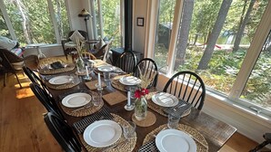 Un espace repas avec une longue table en bois, dressée pour six personnes, entourée de chaises, et de grandes fenêtres donnant sur la forêt.