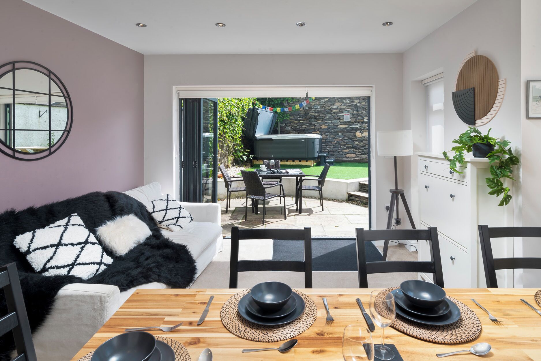 Un comedor con una mesa lista para cuatro personas, un sofá con una manta blanco y negro, y una pared de piedra al fondo.