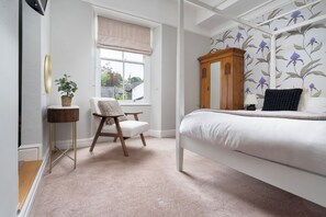 A bedroom with a bed, a chair, a window, a wooden cabinet, and a wall with floral wallpaper. - Rosemount Boutique (Windermere)