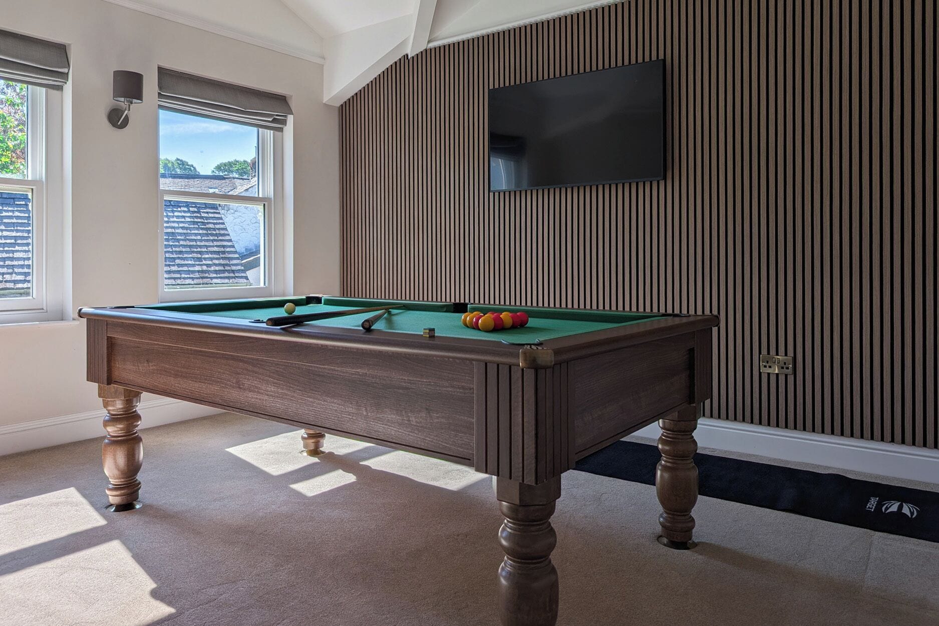 Una mesa de billar con bolas, un televisor montado en la pared y una ventana con persianas.