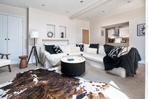 Ein modernes Wohnzimmer mit einem weißen Sofa, einem runden Couchtisch, einem Kuhfellteppich und einem durch einen offenen Durchgang einsehbarer Küchenbereich.