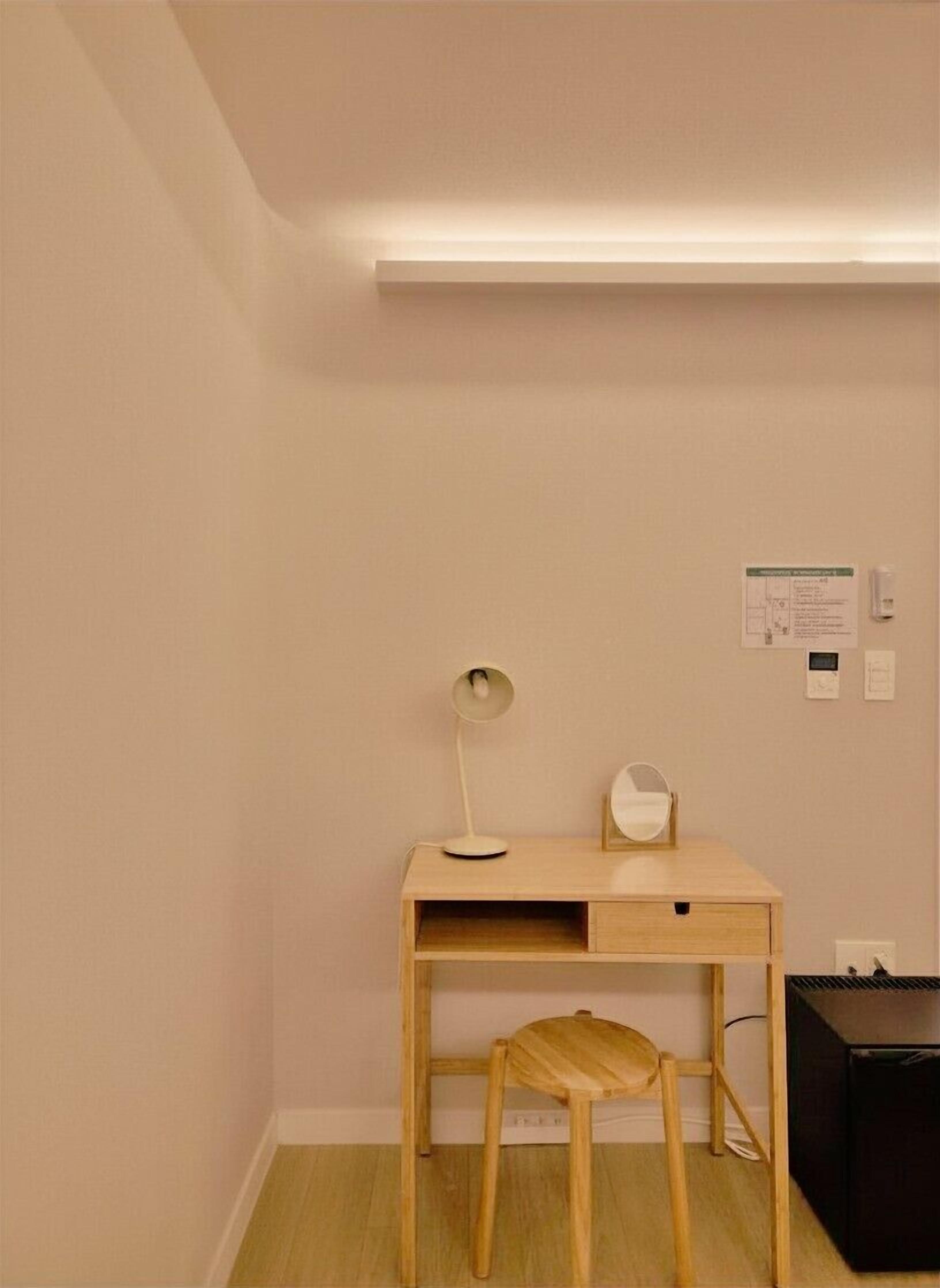 A small, simple room with a wooden desk, a stool, a desk lamp, a small mirror, and a black speaker.