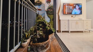 Un moderno vestíbulo de hotel con una gran pantalla de televisión mostrando un programa, un mural decorativo y un balcón con plantas en macetas.