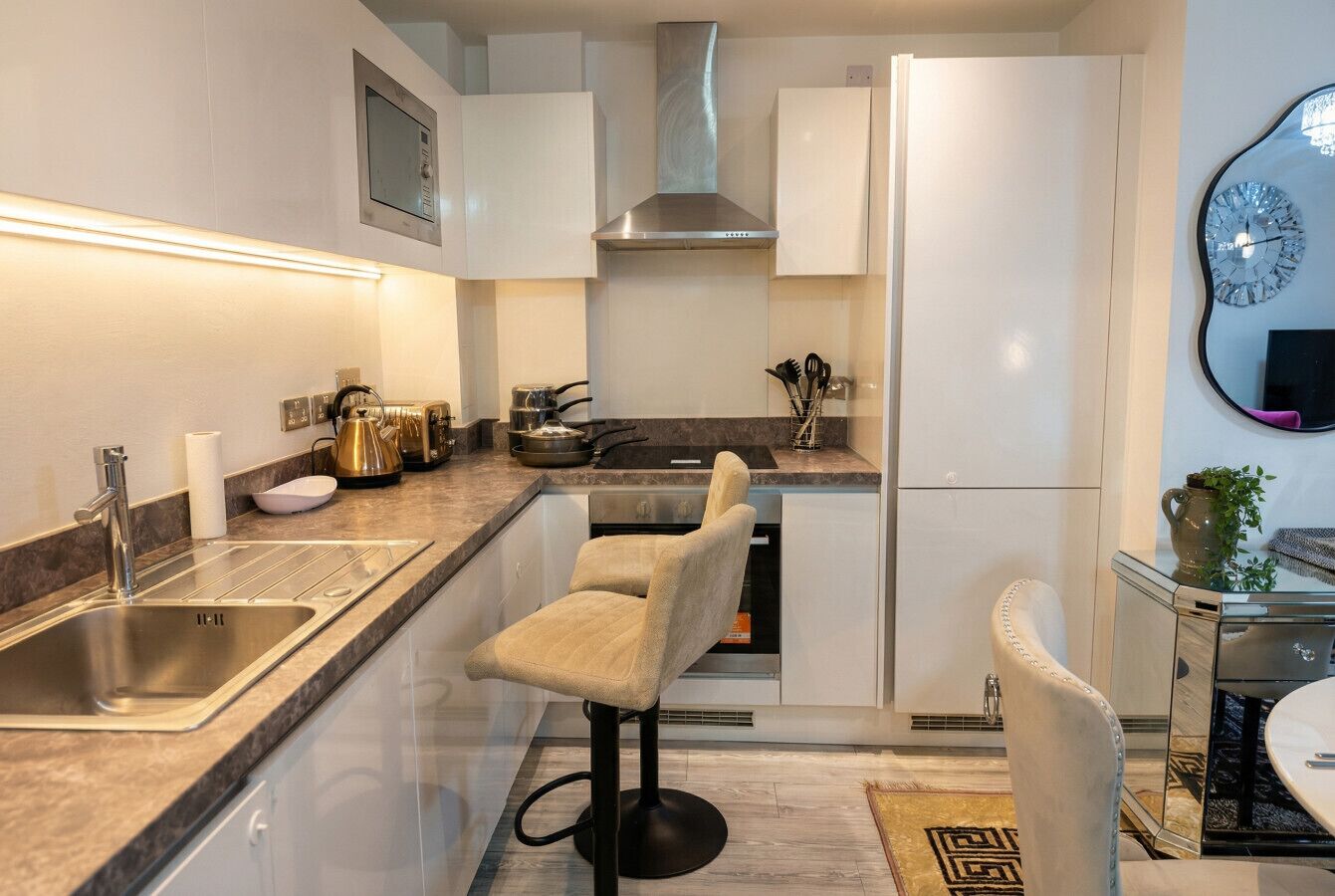 Una cocina moderna con un lavamanos de acero inoxidable, una estufa a gas y un horno empotrado. Hay dos taburetes de bar y una mesa de comedor con un jarrón de flores.