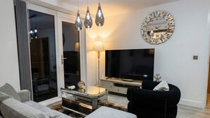 Uma sala de estar moderna com um sofá, uma poltrona preta, uma mesa de centro de vidro, uma televisão e um relógio decorativo.