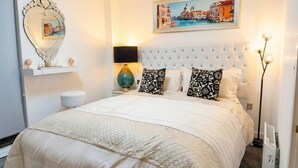 Quarto com cabeceira estofada, cama com roupa de cama branca, espelho decorativo, quadro com vista urbana e abajur.