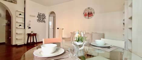 Un comedor con mesa de vidrio, platos blancos y un vaso con una planta. Hay un reloj de pared y un adorno mural.