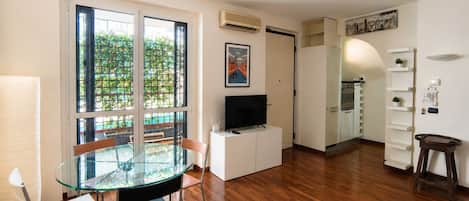 Una sala de estar moderna con una mesa de comedor de vidrio, un televisor de pantalla plana y una zona de cocina con armarios blancos.
