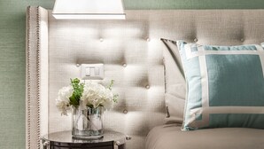 Een slaapkamer met een gestoffeerd hoofdeinde, een nachtkastje met een lamp, een vaas met bloemen en lijstwerken aan de muur.