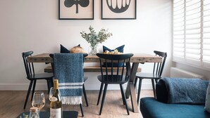Een eetgedeelte met een houten tafel, zwarte stoelen, een blauwe stoel met een herringbon patroon, een blauwe bank, een vaas met bloemen, een fles wijn en twee kunstwerken in lijsten aan de muur.