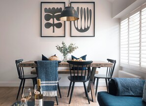 Ein Essbereich mit Holztisch, schwarzen Stühlen, einem blau karierten Sessel, einer blauen Couch, einer Vase mit Blumen, einer Weinflasche und zwei gerahmten Kunstwerken an der Wand.