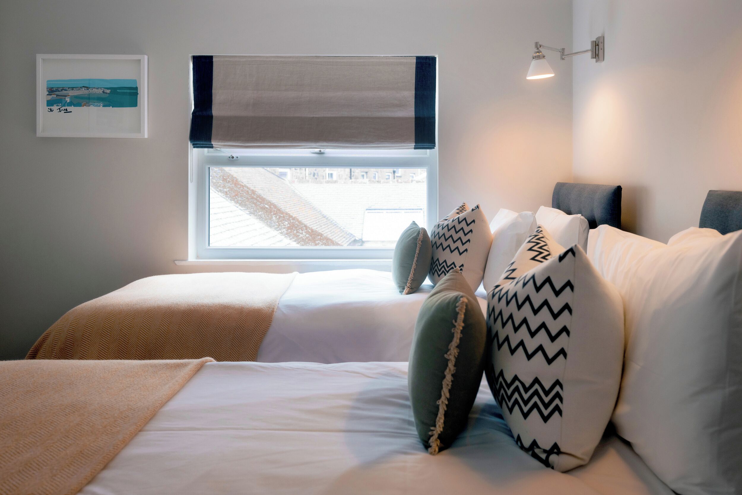 A bedroom with a bed, pillows, a window with a view, a wall-mounted lamp, and a framed picture.