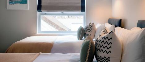 A bedroom with a bed, pillows, a window with a view, a wall-mounted lamp, and a framed picture.