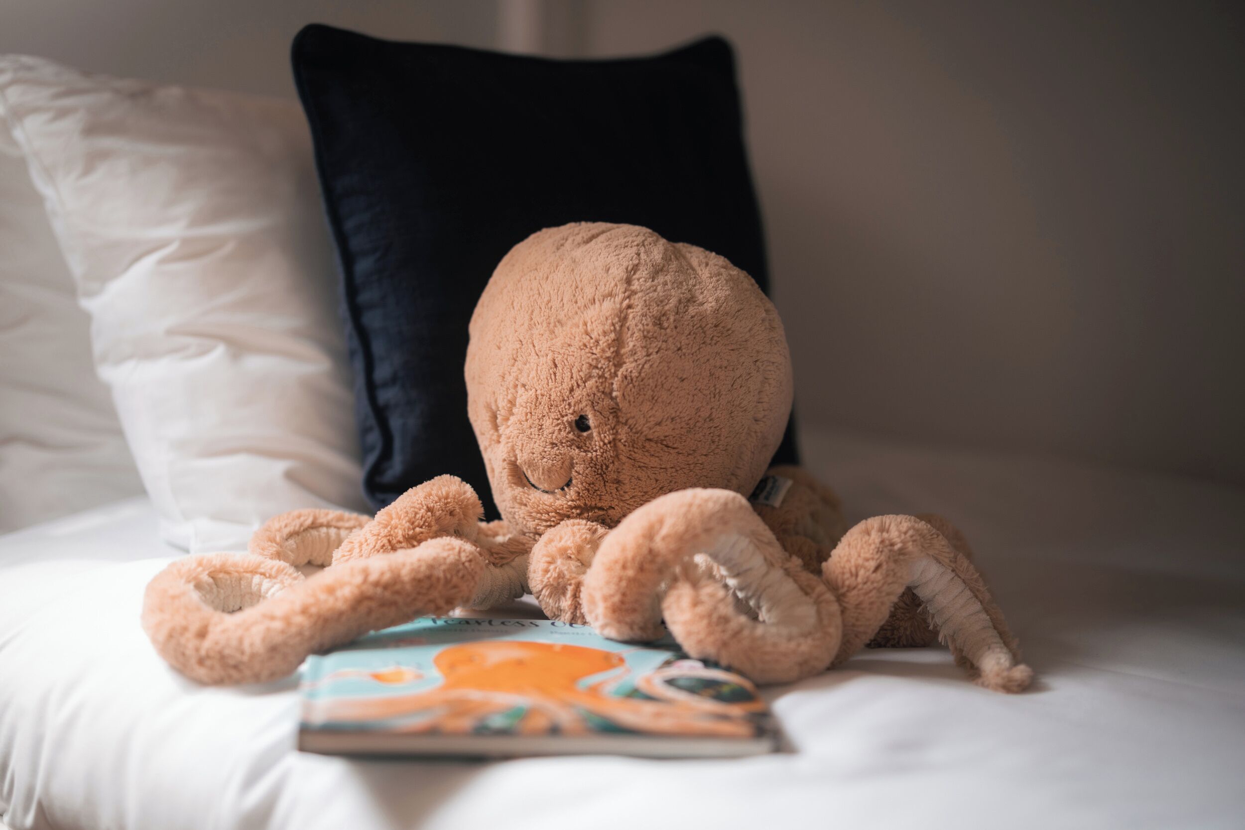 A plush toy on a bed with pillows, a book, and a dark pillow in the background.