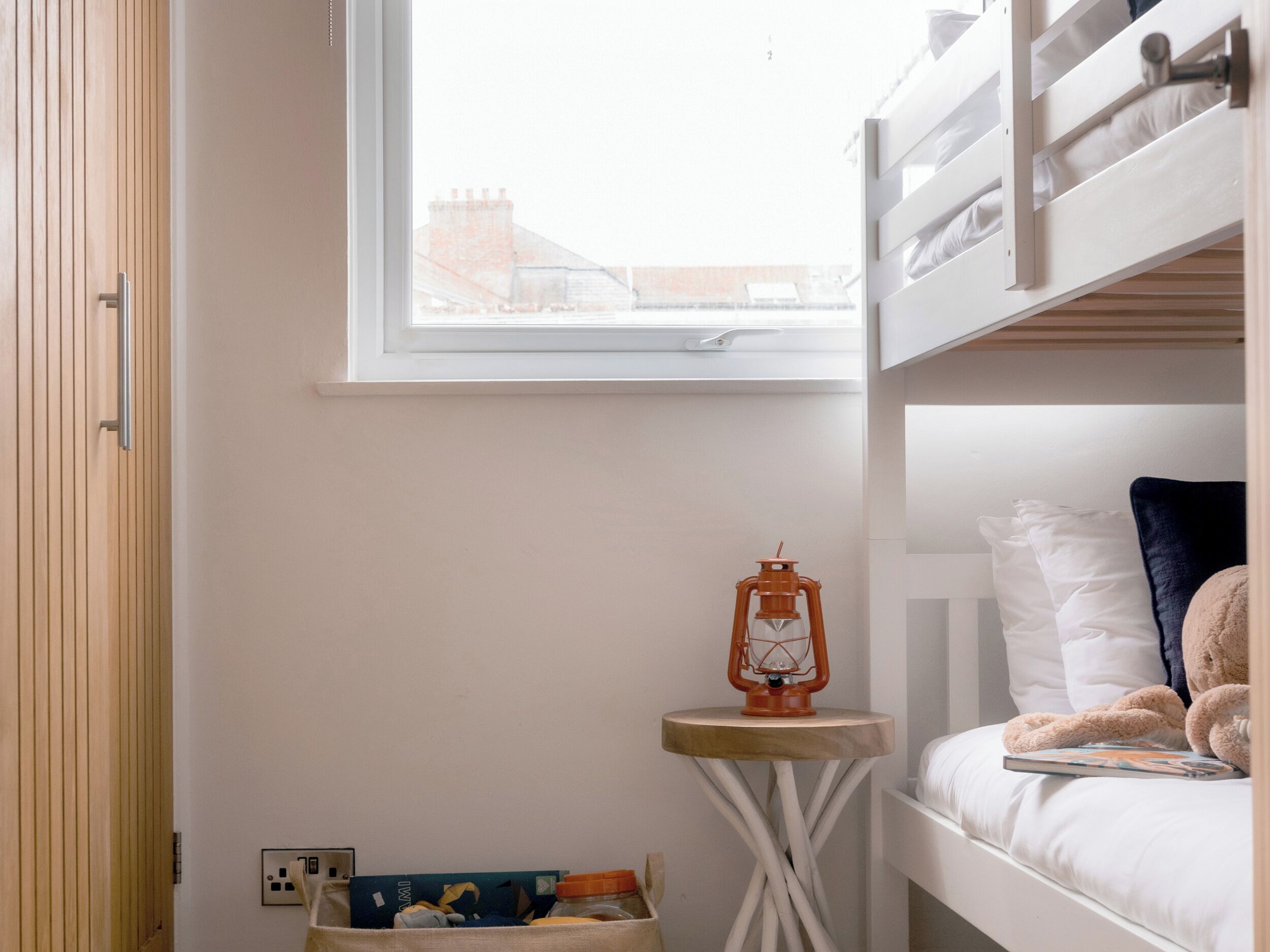 Une chambre avec un lit superposé, une fenêtre, une petite table avec une lanterne et une poubelle.