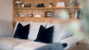 Een lichtkleurige bank met donkere kussens, een houten plank met boeken en decoratieve objecten, en een onscherpe voorgrond.
