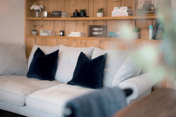 A light-colored sofa with dark cushions, a wooden shelf with books and decorative items, and a blurred foreground. - 28 Piazza (St Ives)