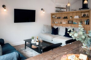 A modern living room with a flat-screen TV, a wooden bookshelf, a grey sofa, a coffee table, and a dining table with a vase of flowers. - 28 Piazza (St Ives)