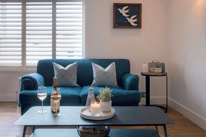 Ein Wohnzimmer mit einem blauen Sofa, einem kleinen Tisch mit Kerze, einer Flasche und einem Glas Weißwein.