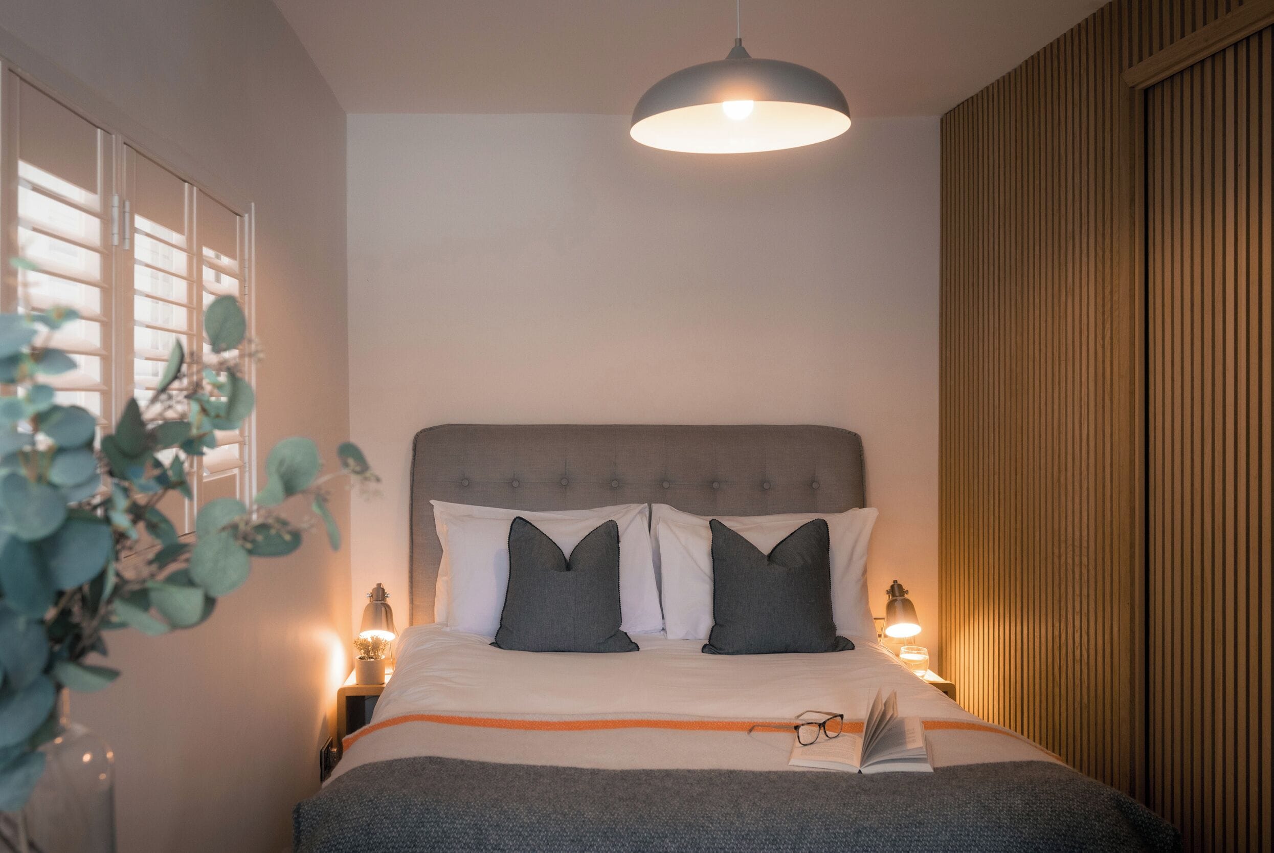 Una cama bien hecha con un cabecero gris, dos almohadas, una lámpara de noche y un libro sobre la cama.