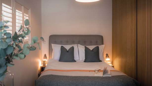 Een net opgemaakt bed met een grijs hoofdeinde, twee kussens, een bedlampje en een boek op het bed.