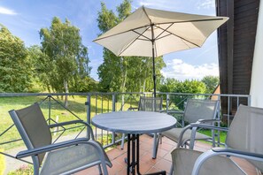 Eine Terrasse mit rundem Tisch, vier Stühlen, einem Sonnenschirm und Blick auf eine grüne Rasenfläche mit Bäumen.