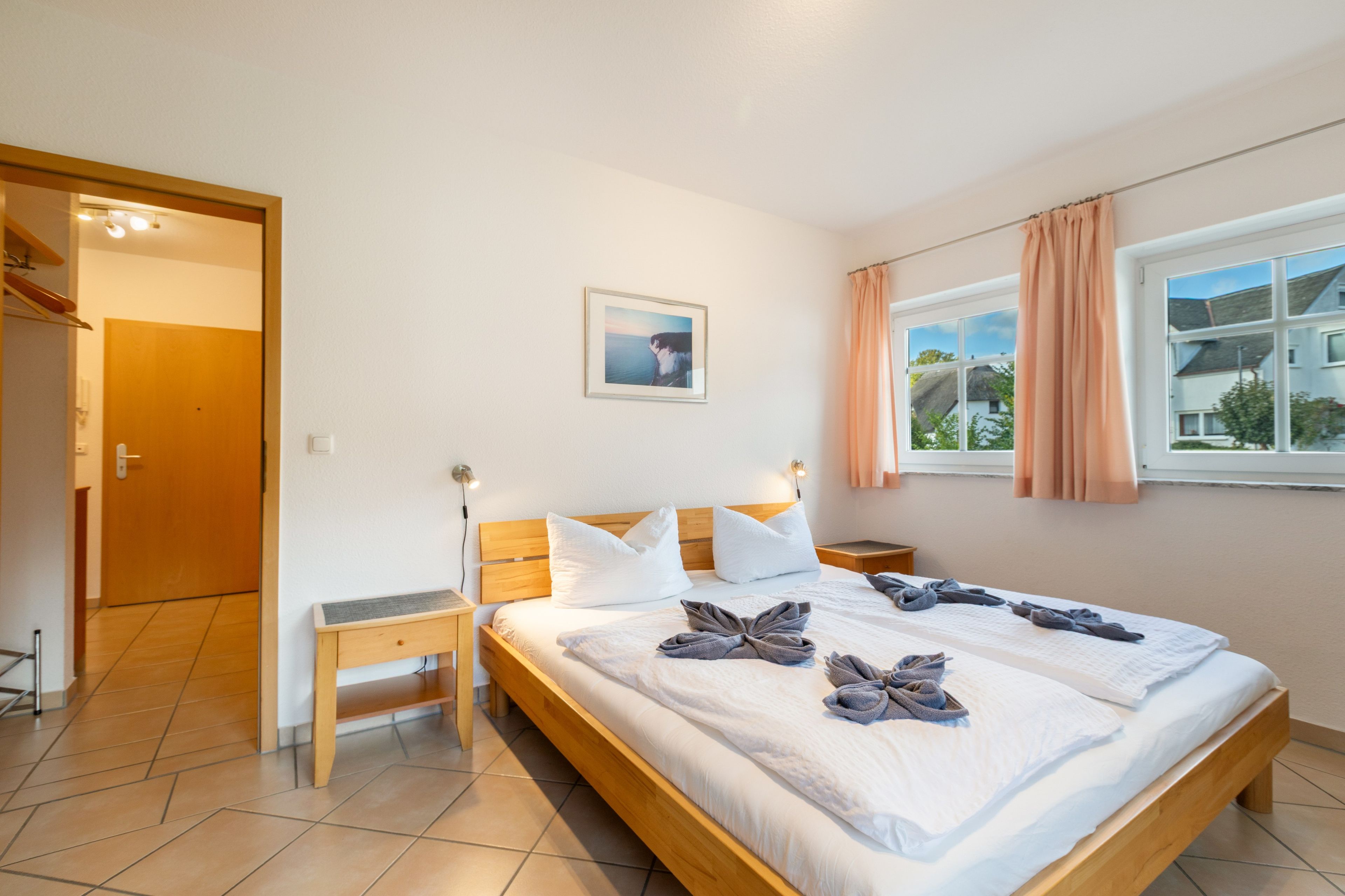 A hotel room with a wooden bed, a bedside table, a nightstand, a window with curtains, and a framed picture on the wall.