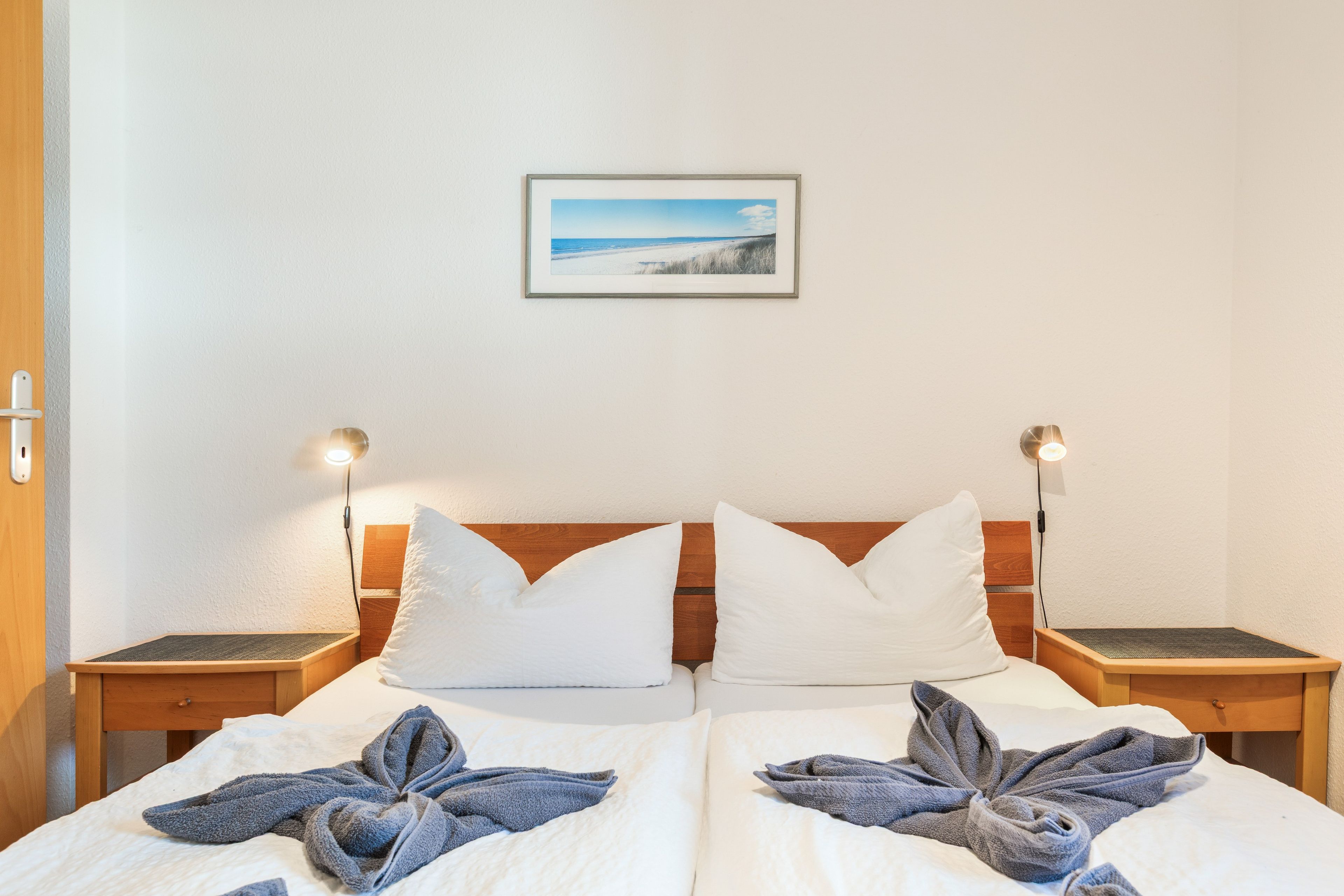 Ein Doppelbett mit weißer Bettwäsche und zwei Kissen, ein Nachttisch mit Lampe sowie ein Bilderrahmen mit einem Strandbild an der Wand.