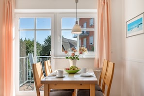 A dining area with a table set for two, a vase with flowers, and a view of a building and trees through large windows. - Dune House with Pool Access in Prime Location (Göhren)