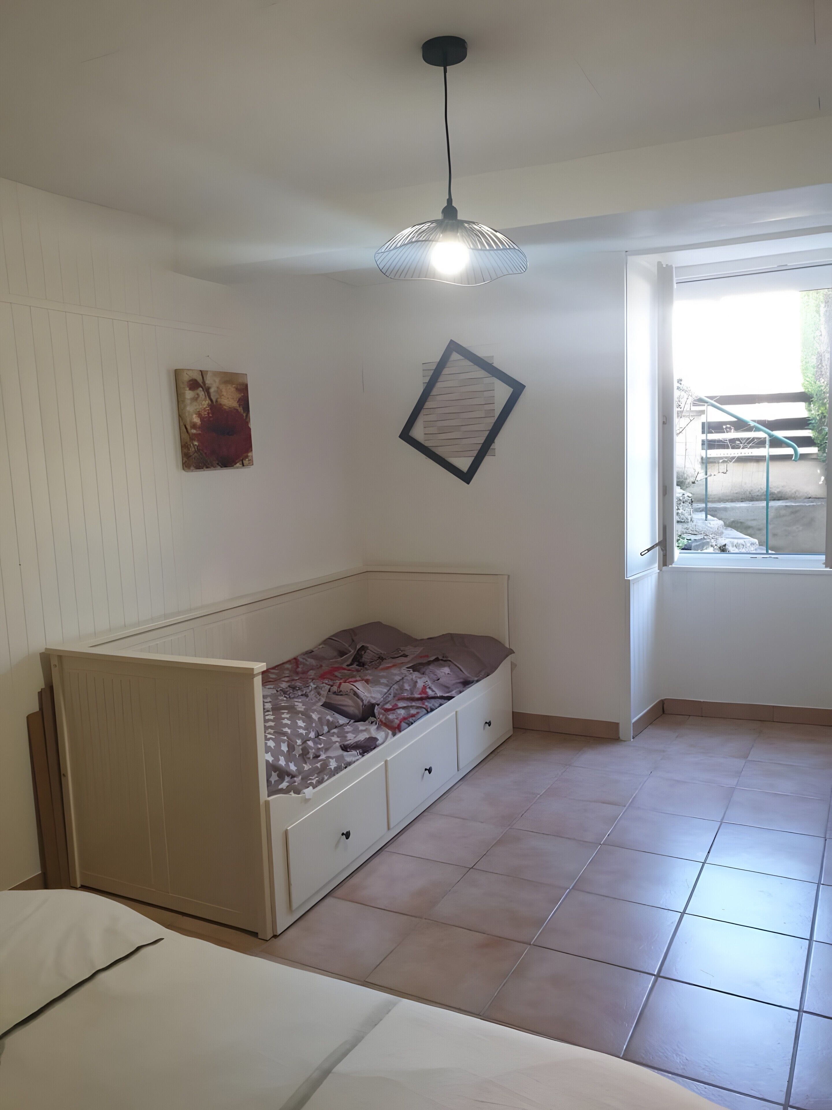 Een kamer met een bed, een hanglamp, een raam en een aan de muur bevestigd schilderij.
