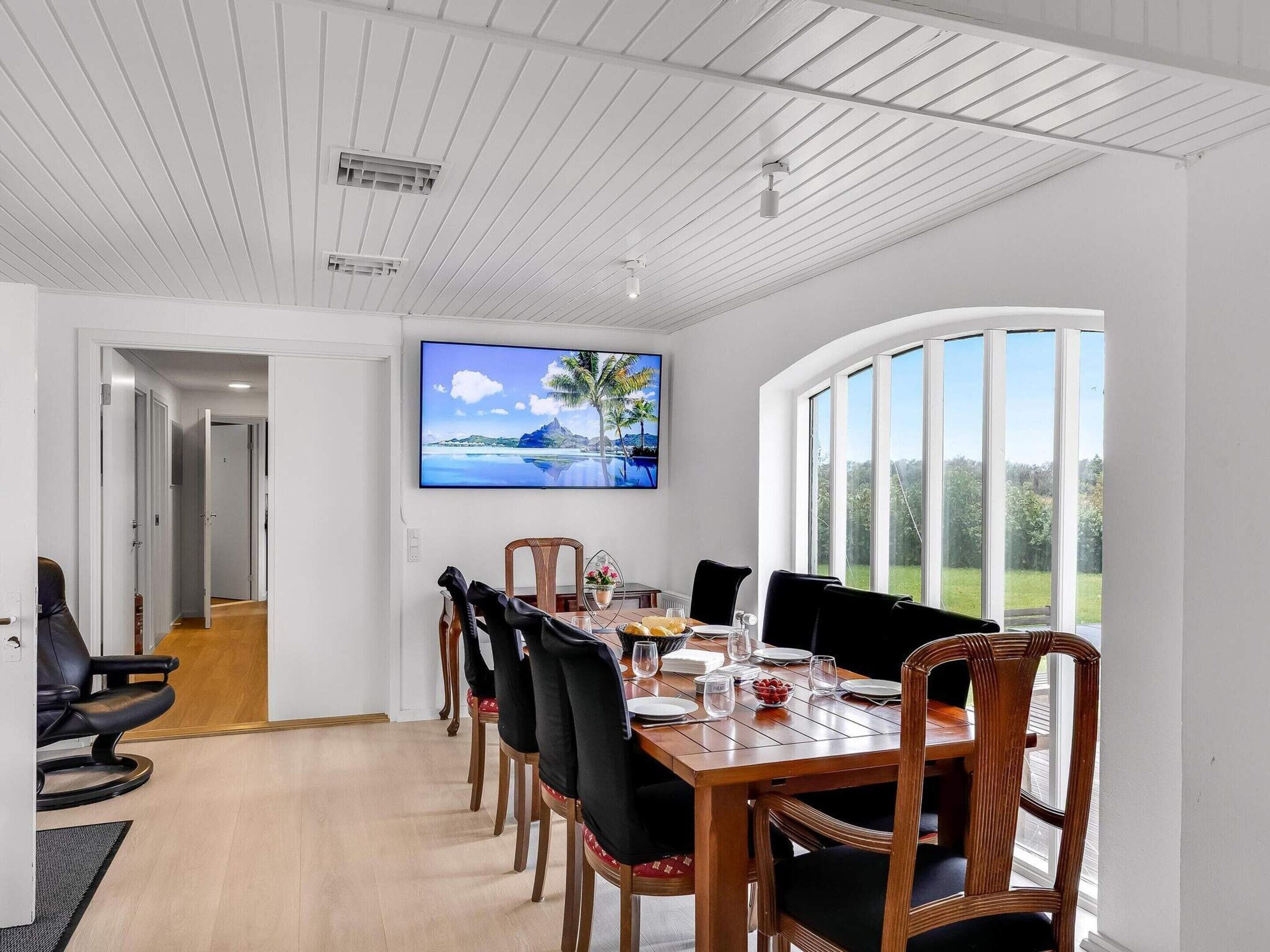 A dining area with a wooden table, chairs, and a television mounted on the wall displaying a scenic image.