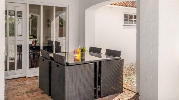 Eine überdachte Terrasse mit Tisch und Stühlen aus Glas, einer weißen Wand und einem kleinen Fenster.