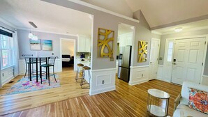 A spacious living area with hardwood floors, a dining table with chairs, a kitchen with white cabinetry, and a hallway leading to bedrooms.
