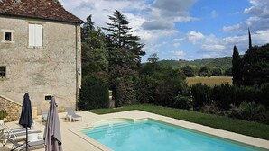 Una piscina con sillas de descanso y sombrillas, un edificio de piedra con tejado de baldosas y un paisaje montañoso al fondo.
