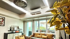 Una sala de estar moderna con sofá, sillas, una mesa de centro y una mesa de comedor. Hay un ventanal con cortinas, un equipo de aire acondicionado y un candelabro.