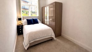 A bedroom with a bed, a nightstand, a wardrobe, and a window with a view of buildings. - Stylish 2-bed Apartment With Patio in Chiswick (London)