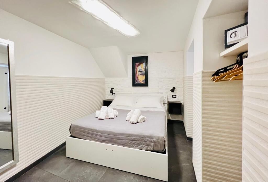 Habitación de hotel con cama, mesita de noche, espejo, un cuadro colgado en la pared y perchero.