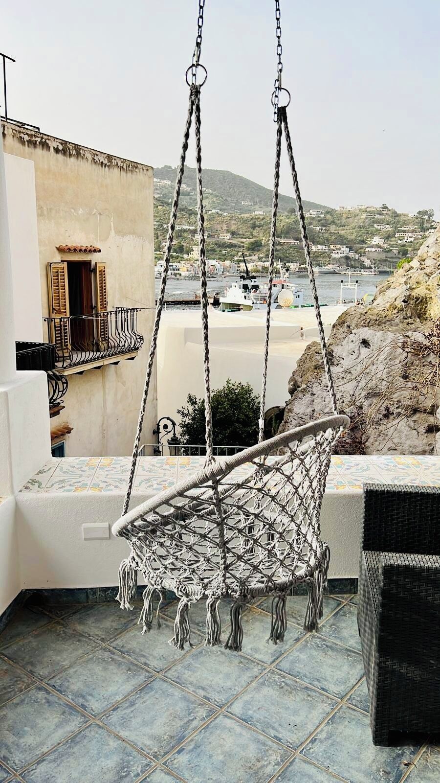 A hammock chair suspended from ceiling chains on a balcony with a view of a coastal town.