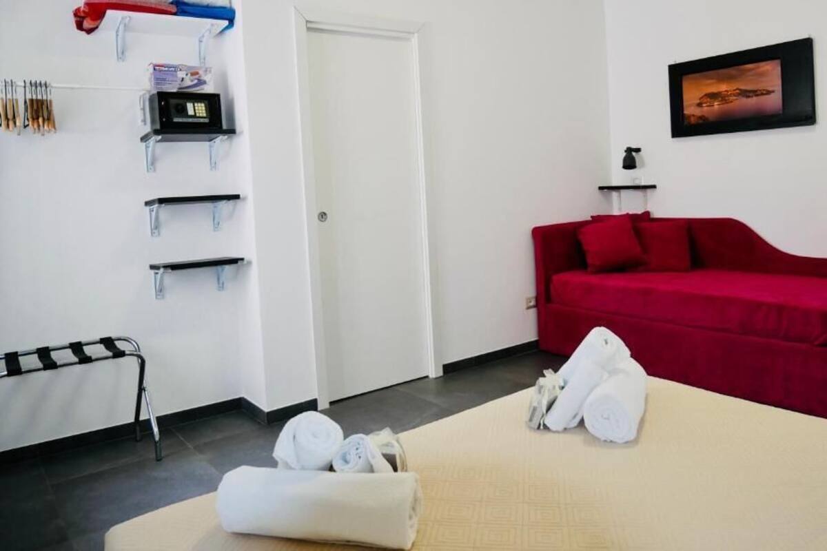 Una habitación de hotel con un sofá rojo, una cama con toallas enrolladas, una puerta blanca, un perchero de pared y un cuadro enmarco.