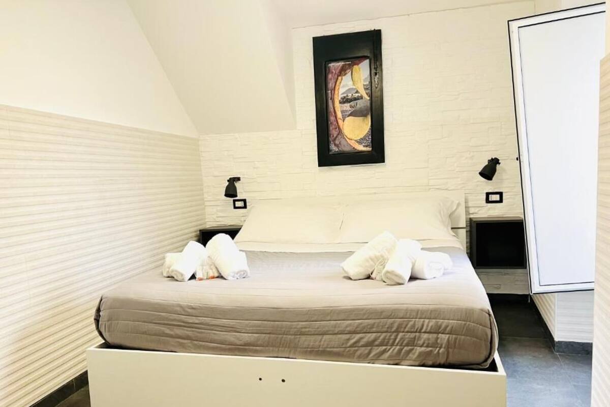 Una habitación de hotel moderna con una cama, paredes blancas, una obra de arte enmarcada y una mesita de noche con una lámpara.